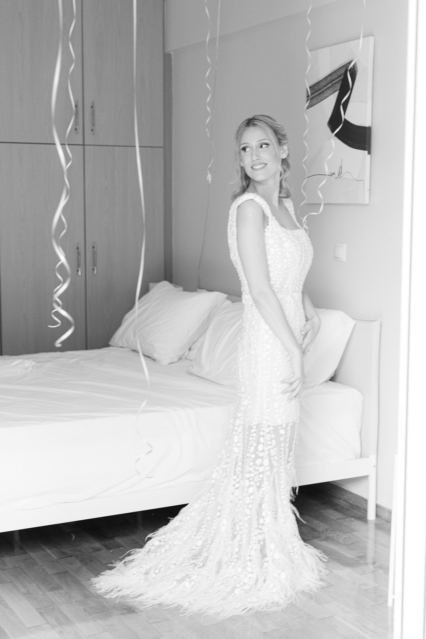 A elegant bride in a form-fitting white lace wedding gown with a mermaid silhouette poses beside a bed with decorative streamers.
