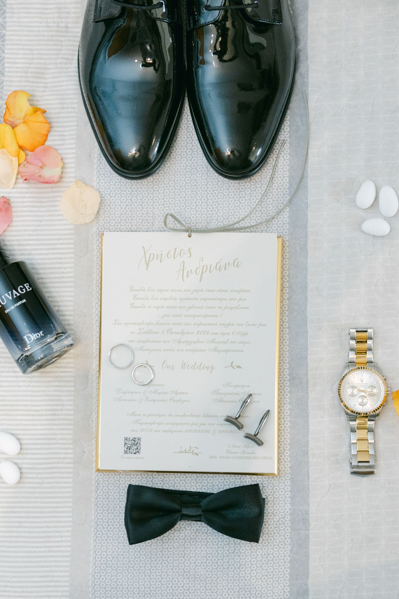 Wedding details flatlay with black dress shoes, invitation, bow tie, gold watch and small decorative items on gray fabric.