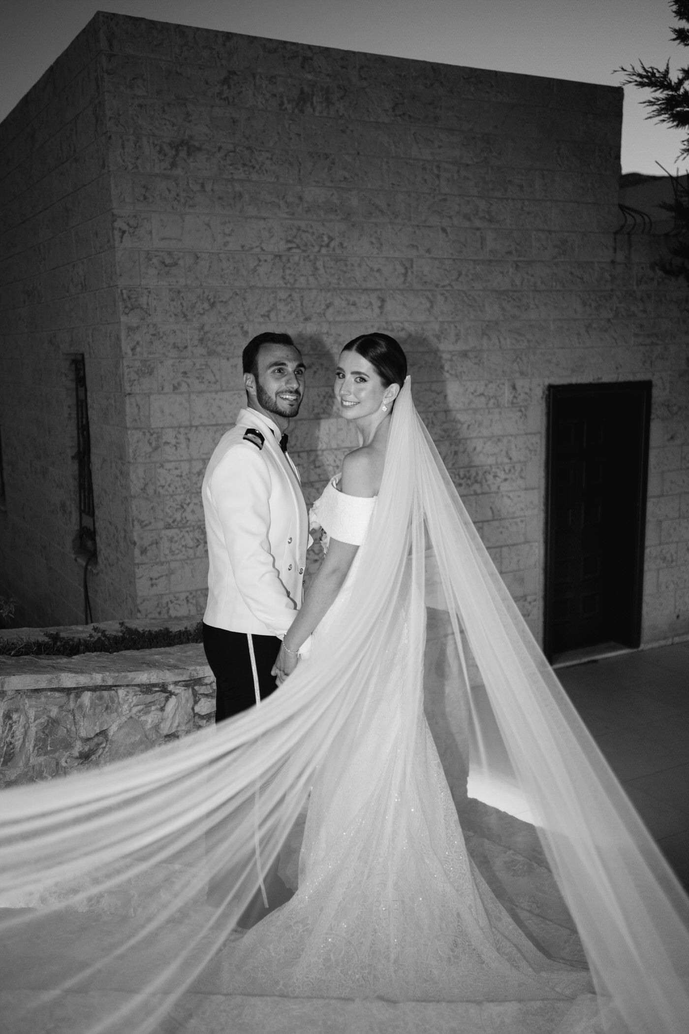 A romantic black and white wedding photo shows newlyweds with a flowing long veil against a stone wall.