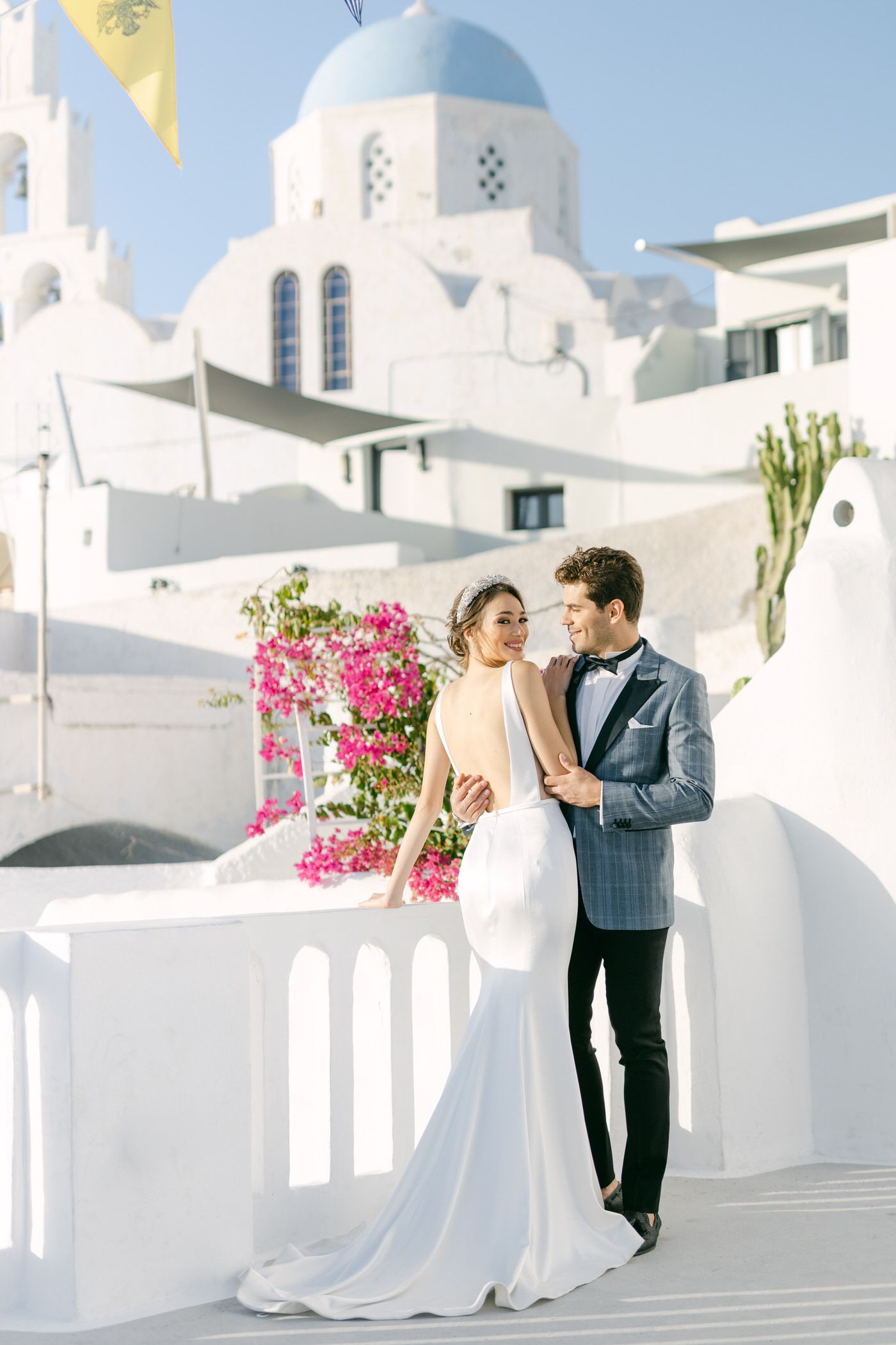 A bride in white gown with long veil and groom in grey suit share a moment while holding pink bouquet in Milos Island