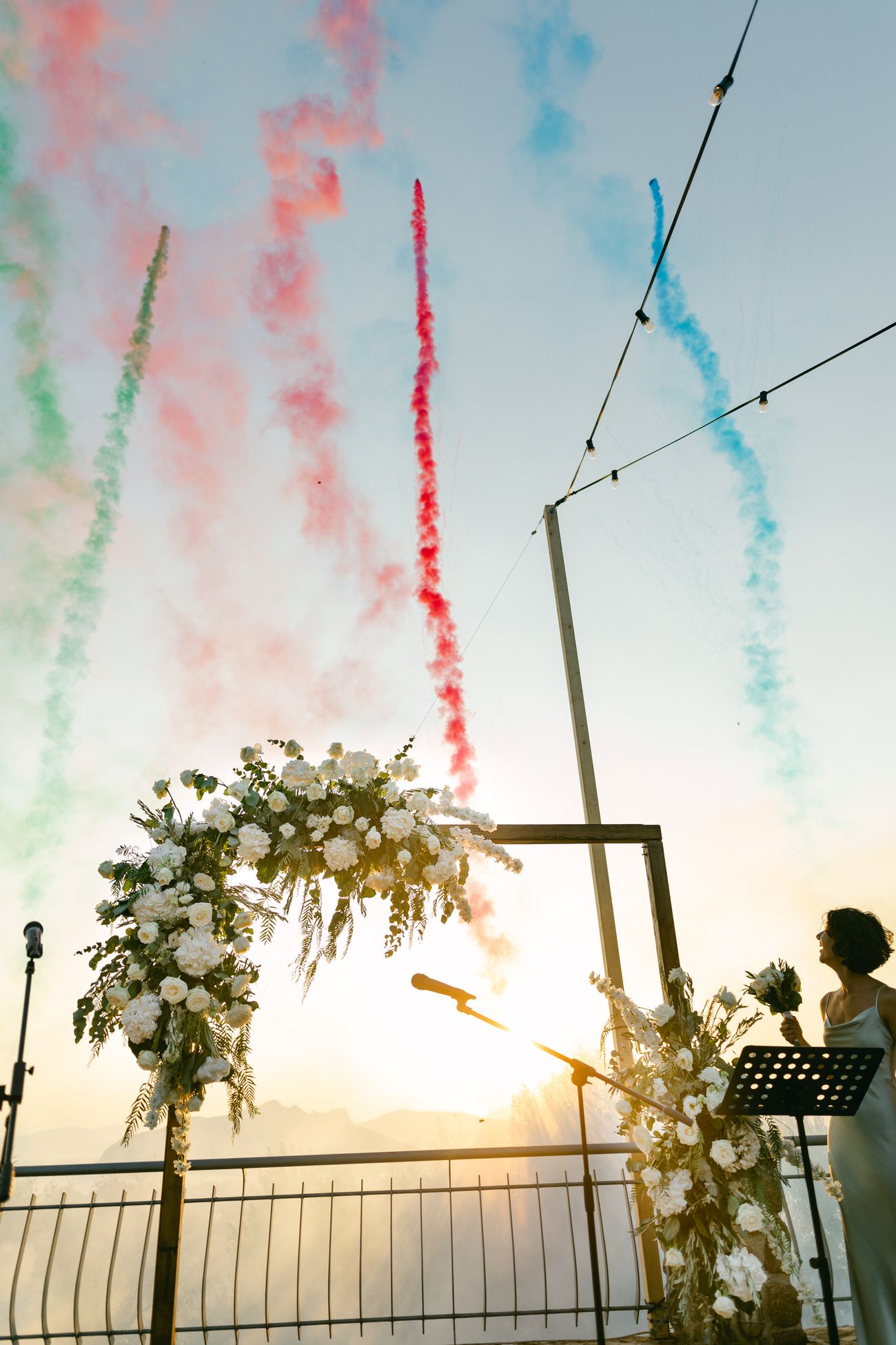 Floral arch silhouetted against sunset sky with colorful smoke trails during outdoor ceremony.