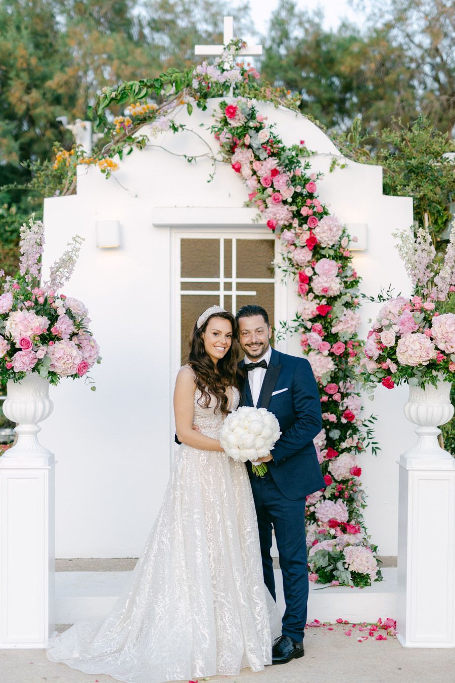 Romantic white wedding arch decorated with pink roses and greenery framing a couple.