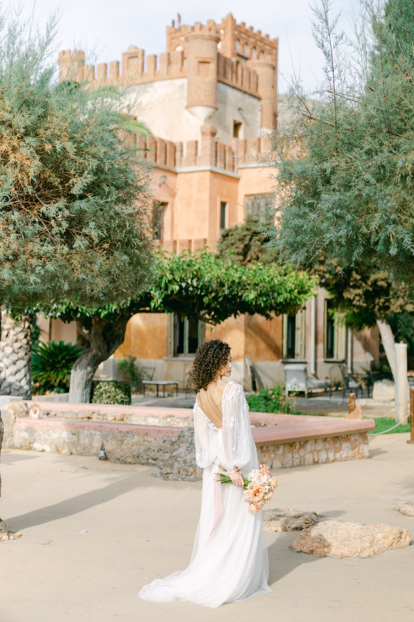 Dreamy castle-like building with tan stone walls and turrets surrounded by lush greenery and garden paths in warm sunlight.