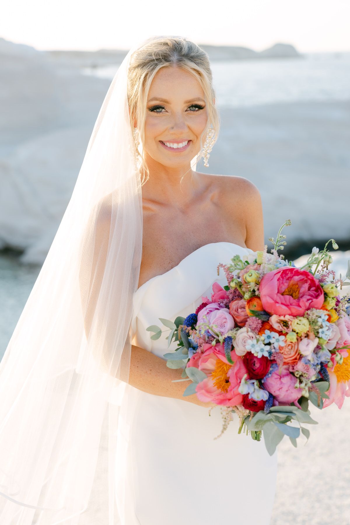 A beautiful bridal portrait featuring a white wedding dress and vibrant pink and coral floral bouquet against a light background.
