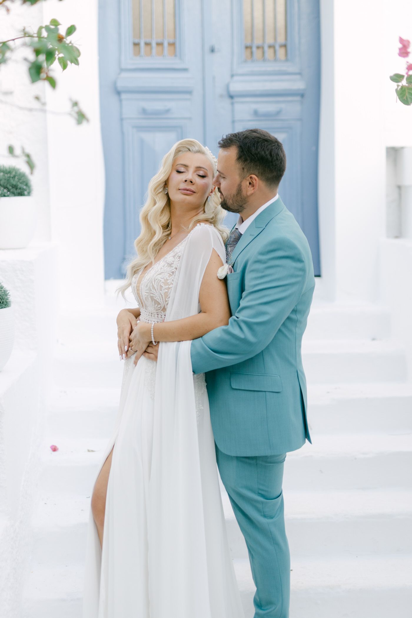 A couple in wedding attire embrace in front of light blue doors with white walls and pink flowers.