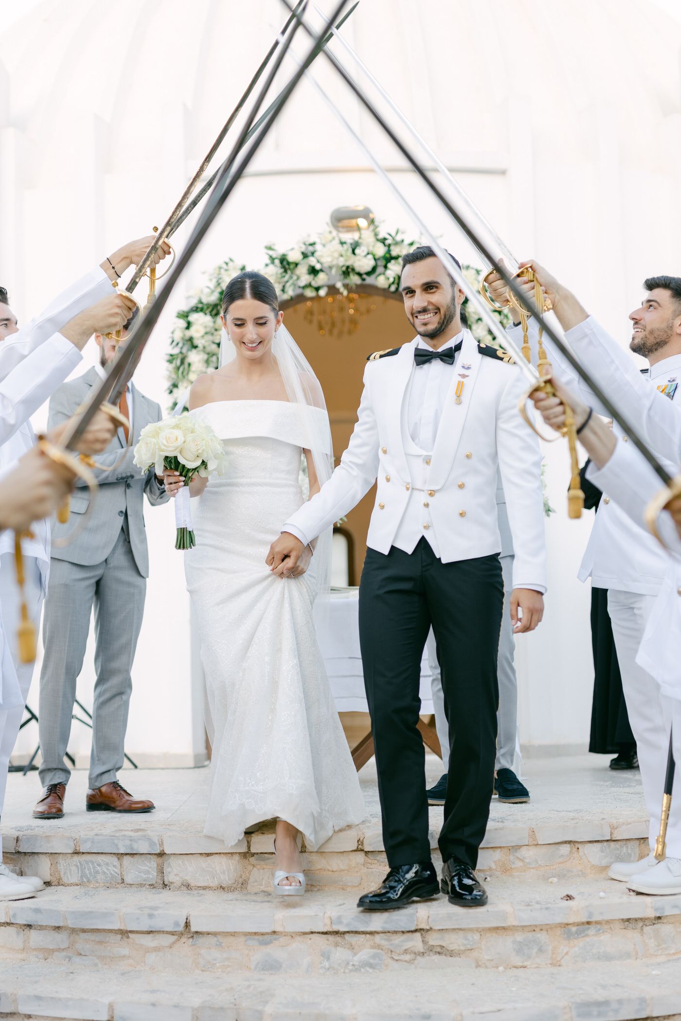 Wedding party creates ceremonial arch with sabers as newlyweds descend white steps under floral decorations.