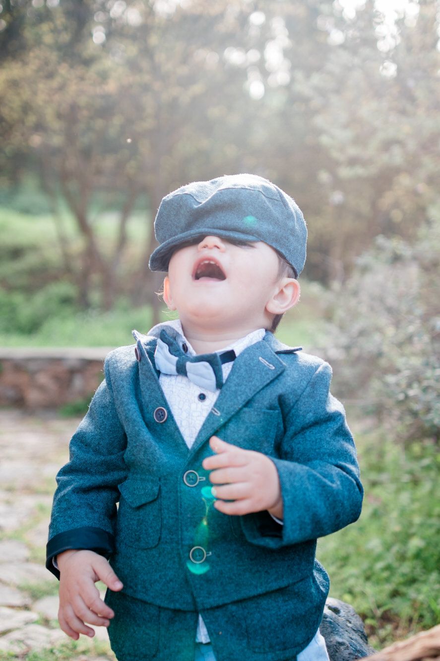 Small child wearing a denim cap and blue blazer laughs joyfully outdoors in a natural setting.