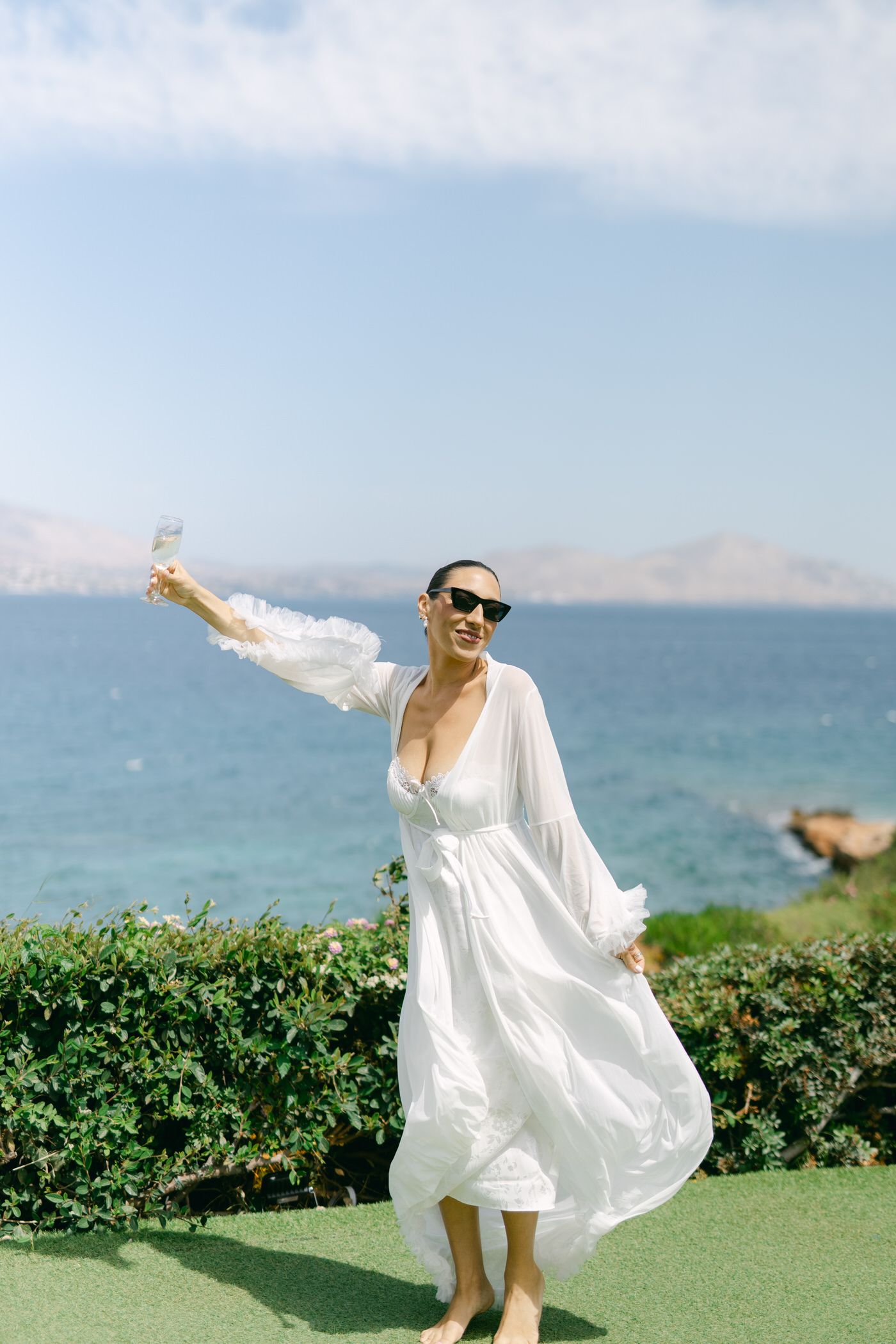 A person in a flowing white dress twirls joyfully on a grassy lawn overlooking the ocean on a sunny day.