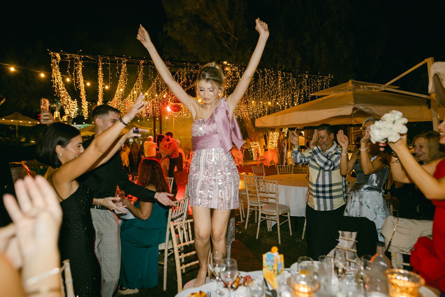 A person in a sparkly dress stands on a chair at an outdoor evening party while others dance and celebrate around them.