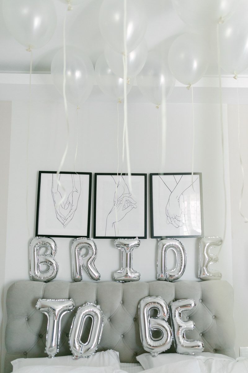 Silver balloon letters spelling 'BRIDE TO BE' displayed on a gray tufted couch with minimalist hand drawings framed above.