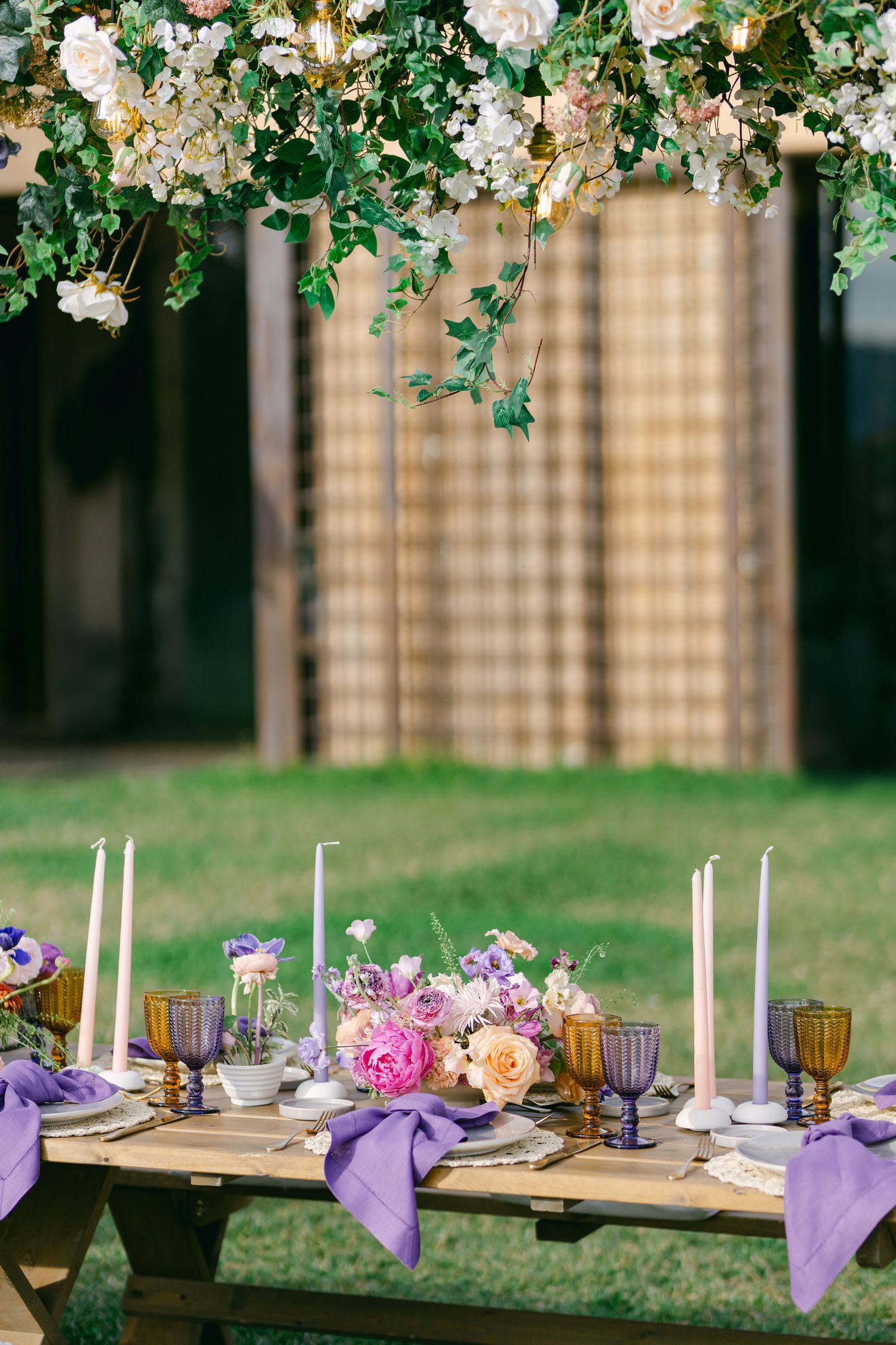 Elegant outdoor table setting with purple linens, white candles and floral arrangements under hanging garden decor.