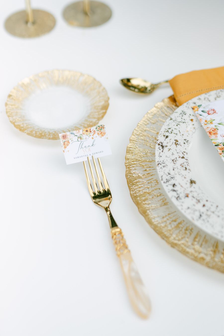 Close up of gold rimmed plate and utensils with delicate place card on white tablecloth.