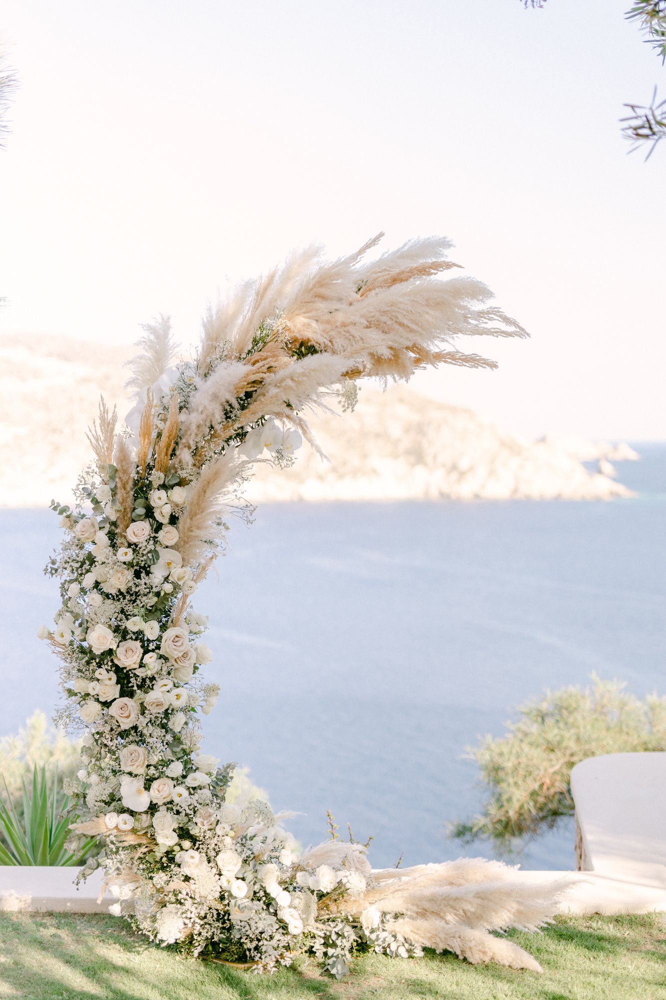 Curved floral wedding arch with white flowers and pampas grass against ocean view.