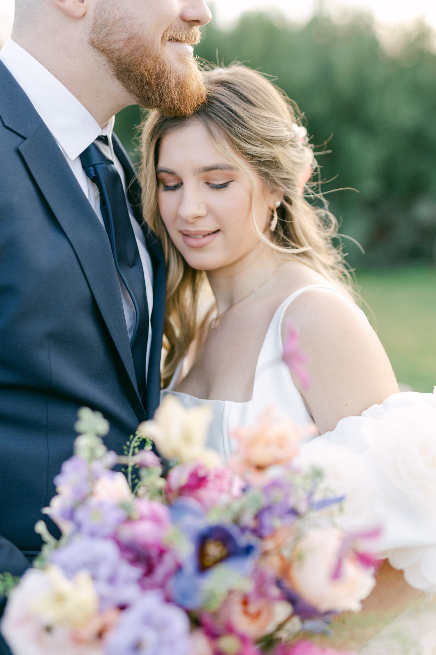 A romantic wedding couple stands close together outdoors with a colorful bouquet of purple and pink flowers.
