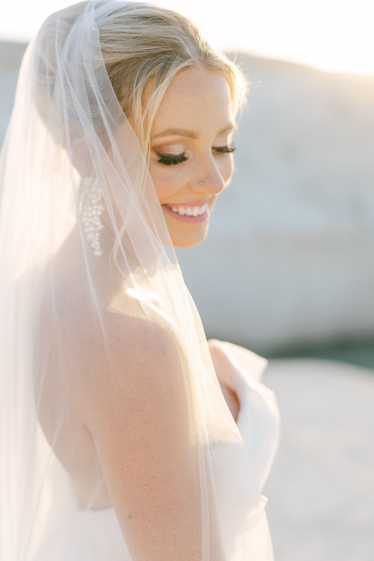A radiant bride wearing a white veil glows in soft sunlight, with delicate makeup and a joyful smile.