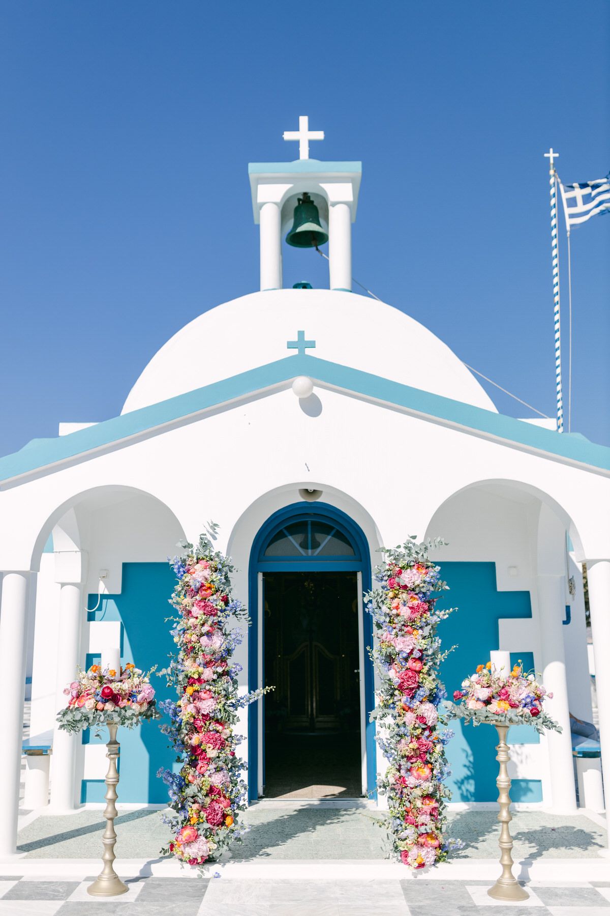 Small white Greek Orthodox church with blue accents and pink floral decorations framing the entrance in Santorini, Greece.