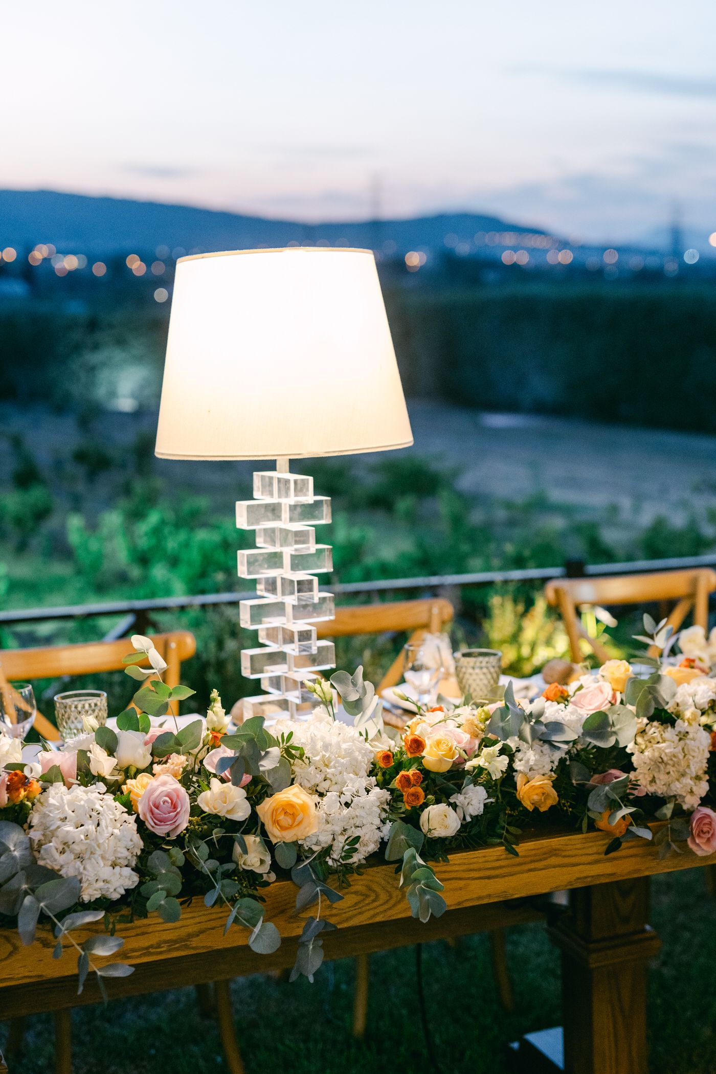 An elegant table lamp illuminates a floral arrangement with white hydrangeas and peach roses against a twilight mountain view.