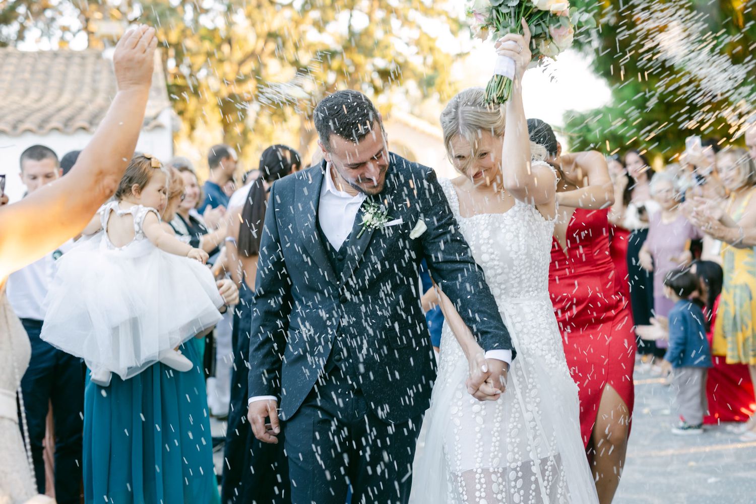 Newlyweds walk through a shower of white confetti while celebrating with wedding guests during an outdoor celebration.