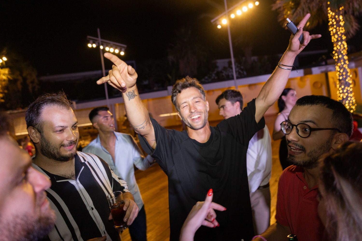 People enjoying a lively nighttime celebration with raised arms and bright lights in the background.