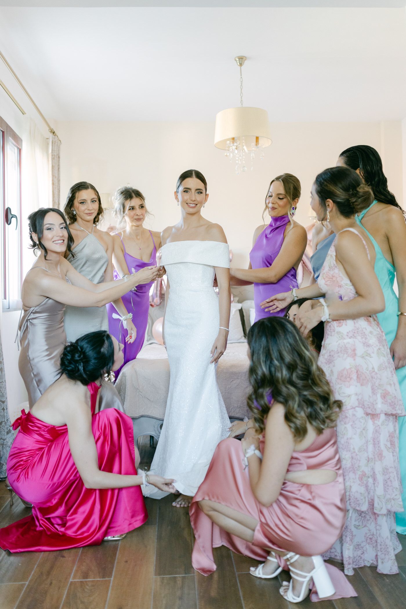 Group helps with a wedding dress fitting in a bright room with pastel colored dresses and elegant decor.