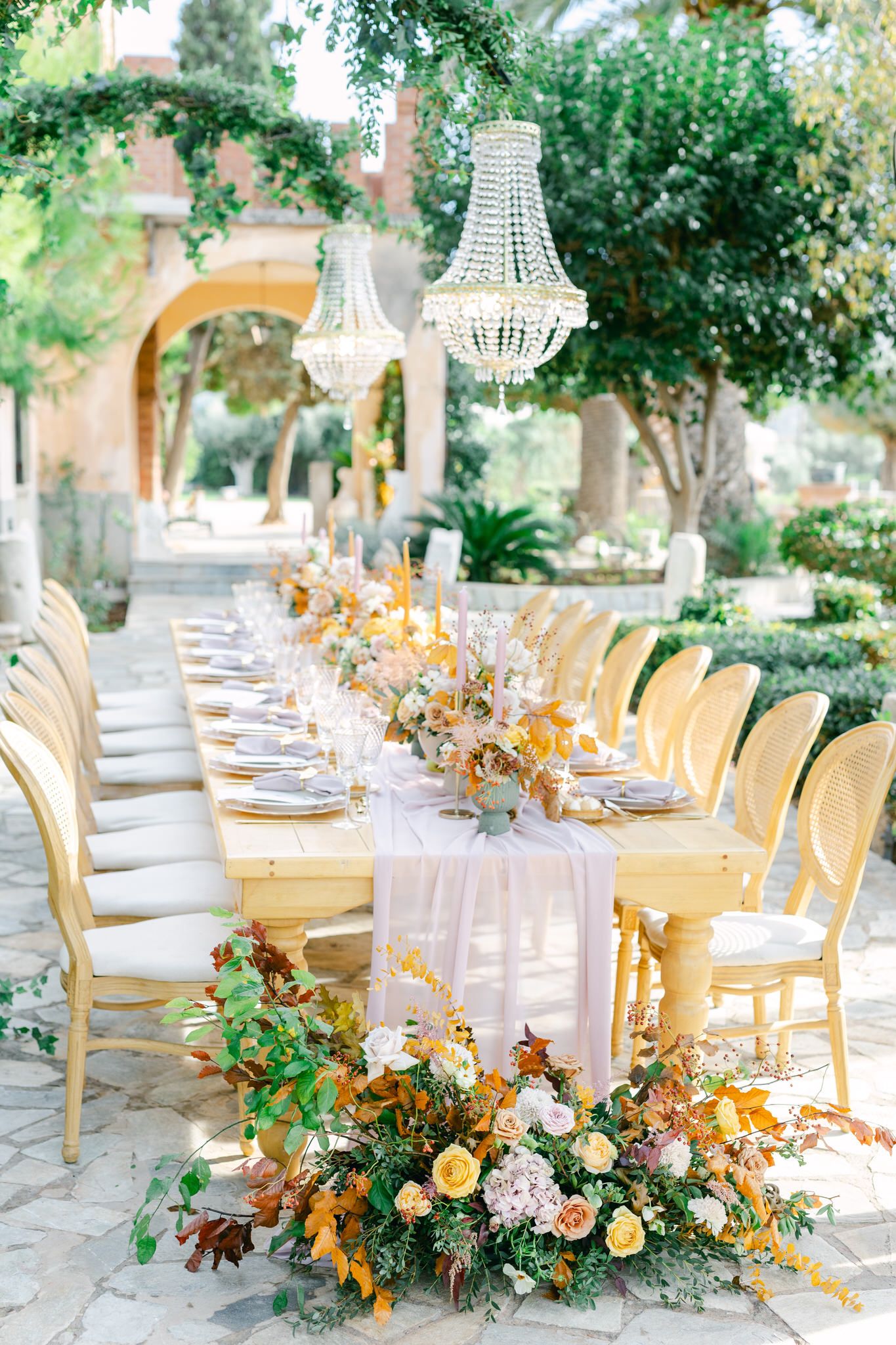 Elegant outdoor wedding reception table with crystal chandeliers and golden chairs along a stone pathway surrounded by greenery.