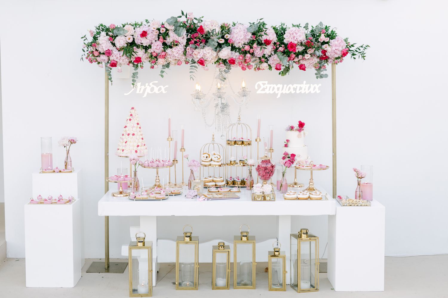 Elegant dessert table with pink flowers, gold lanterns, and white cake stands under a floral arch garland. Ktima 48 baptism Βαπτιση