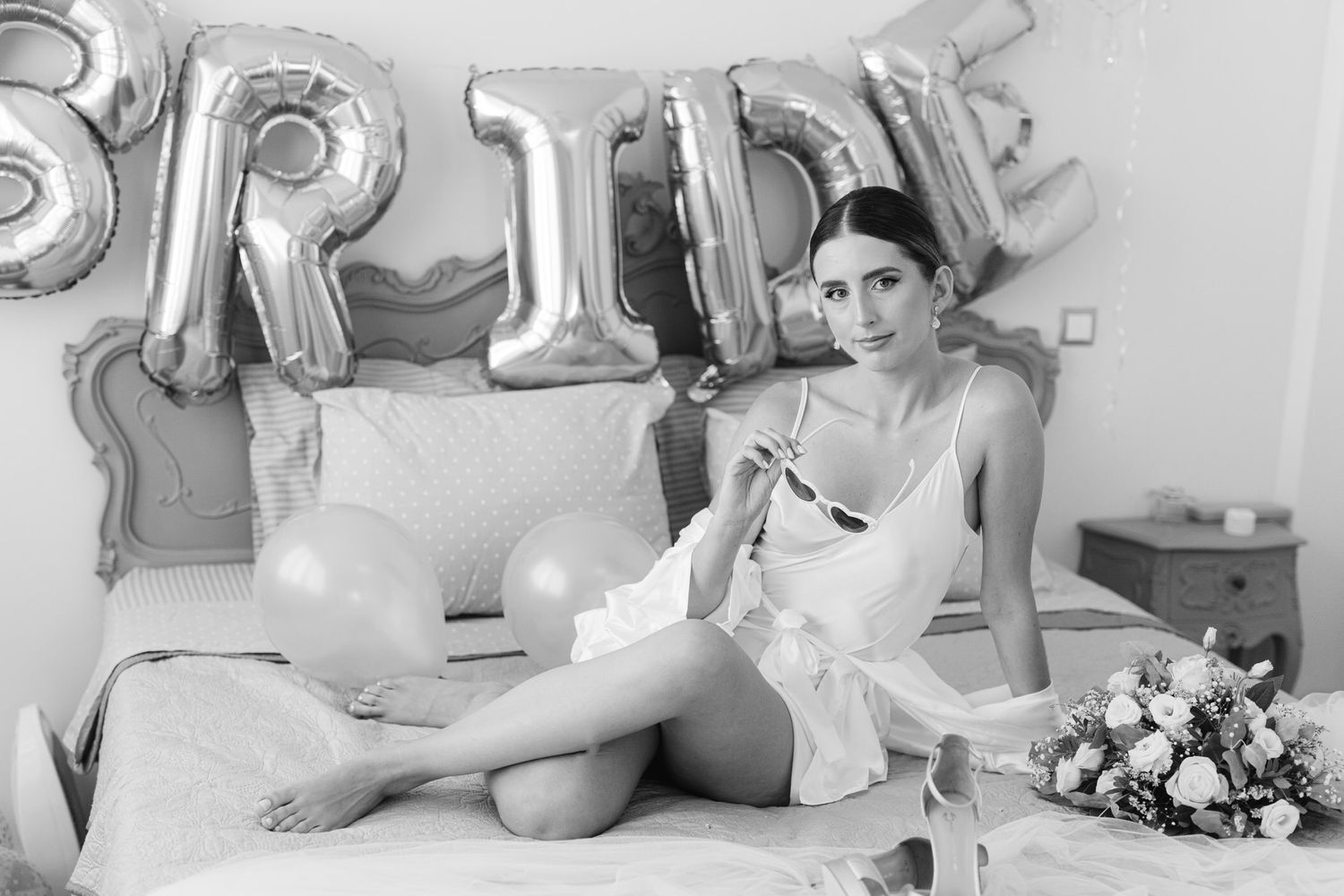 Black and white photo of bride lounging on bed with silver balloon letters and bouquet of flowers.