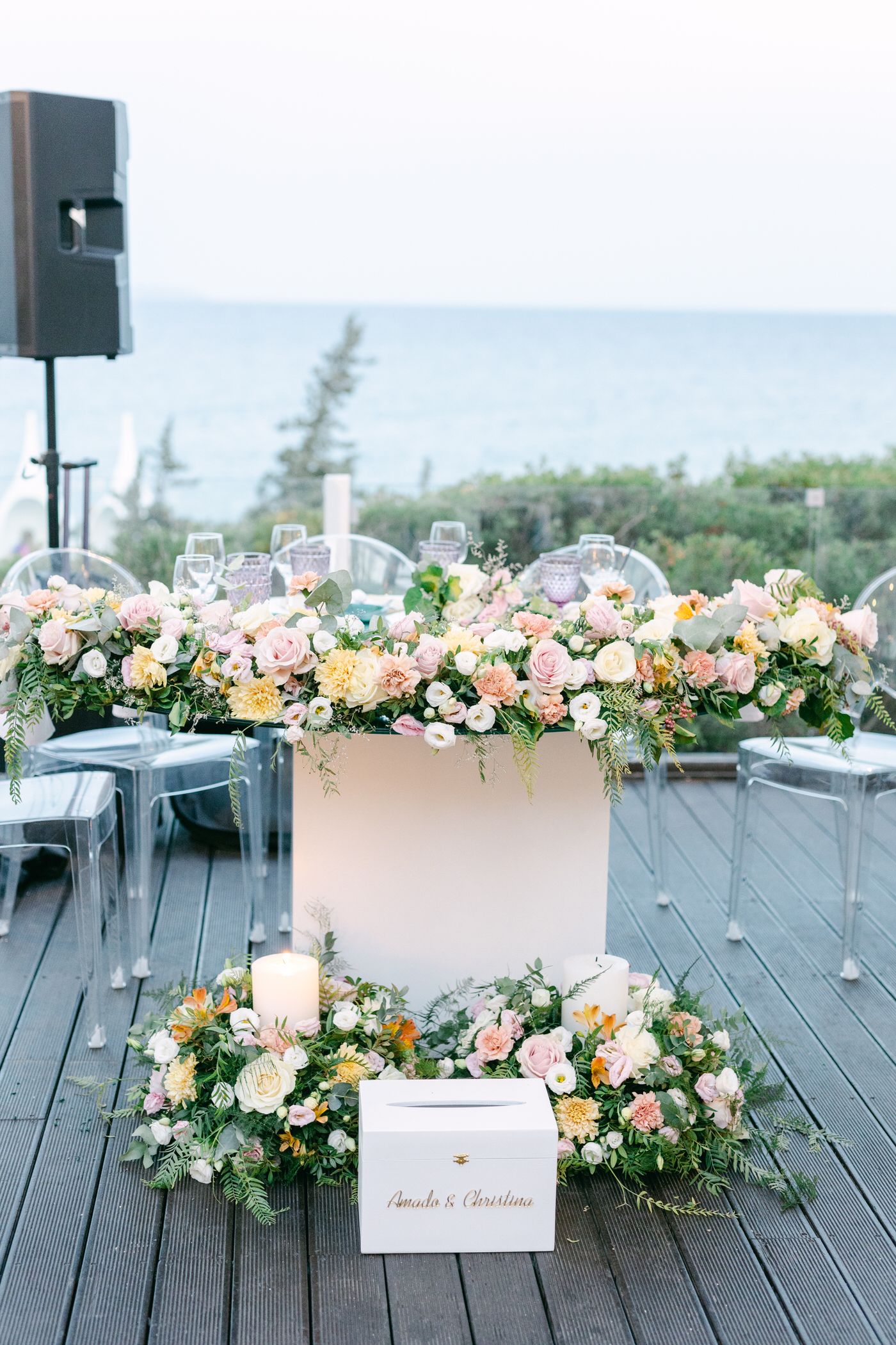 Elegant wedding floral arrangements in peach and pink tones decorate a wooden deck overlooking the ocean at sunset.