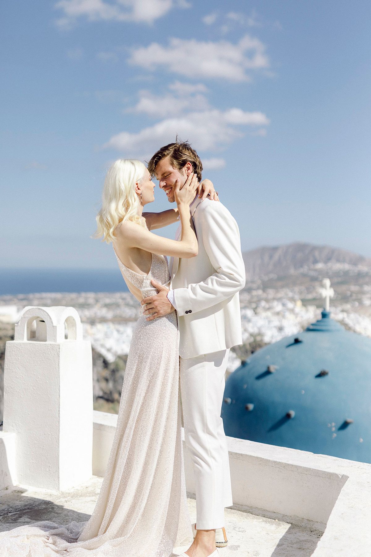 A romantic couple in white attire embraces on a terrace overlooking the blue domes of Santorini, Greece.
