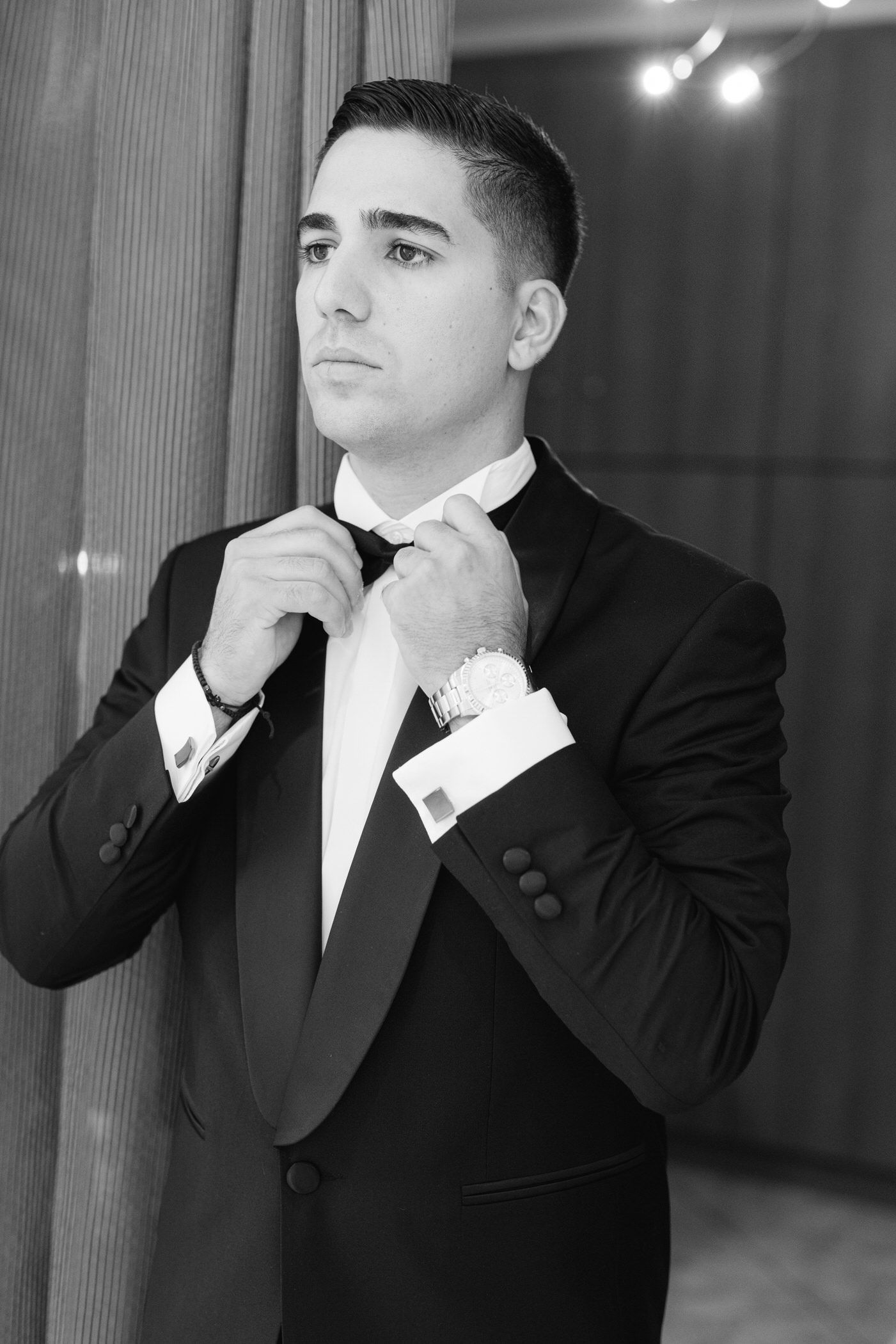 A person in a black tuxedo adjusts their bow tie while standing in a formal setting, captured in black and white.