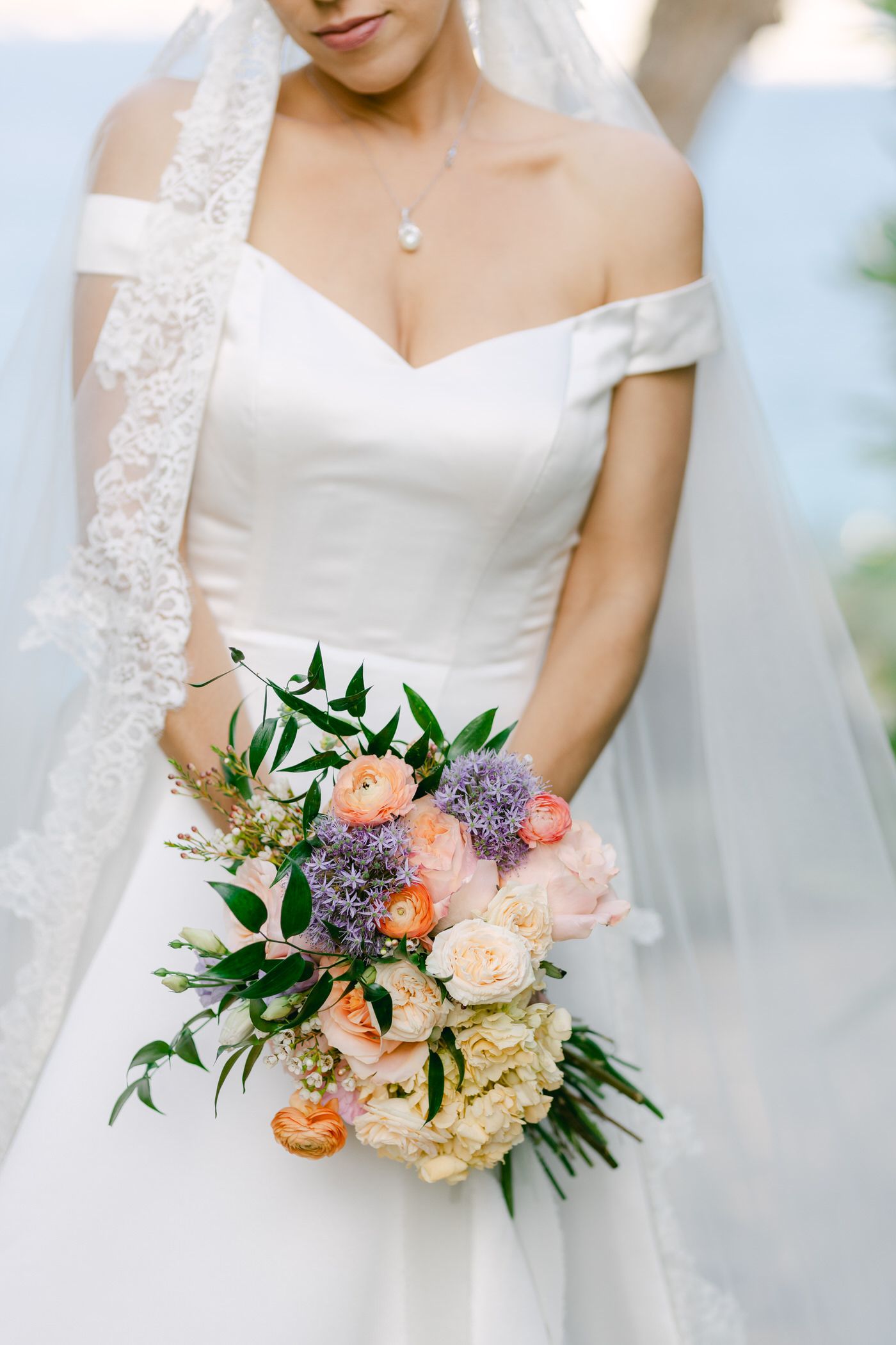 Beautiful bridal bouquet with peach roses, purple allium, and greenery held against an off-shoulder white wedding gown.