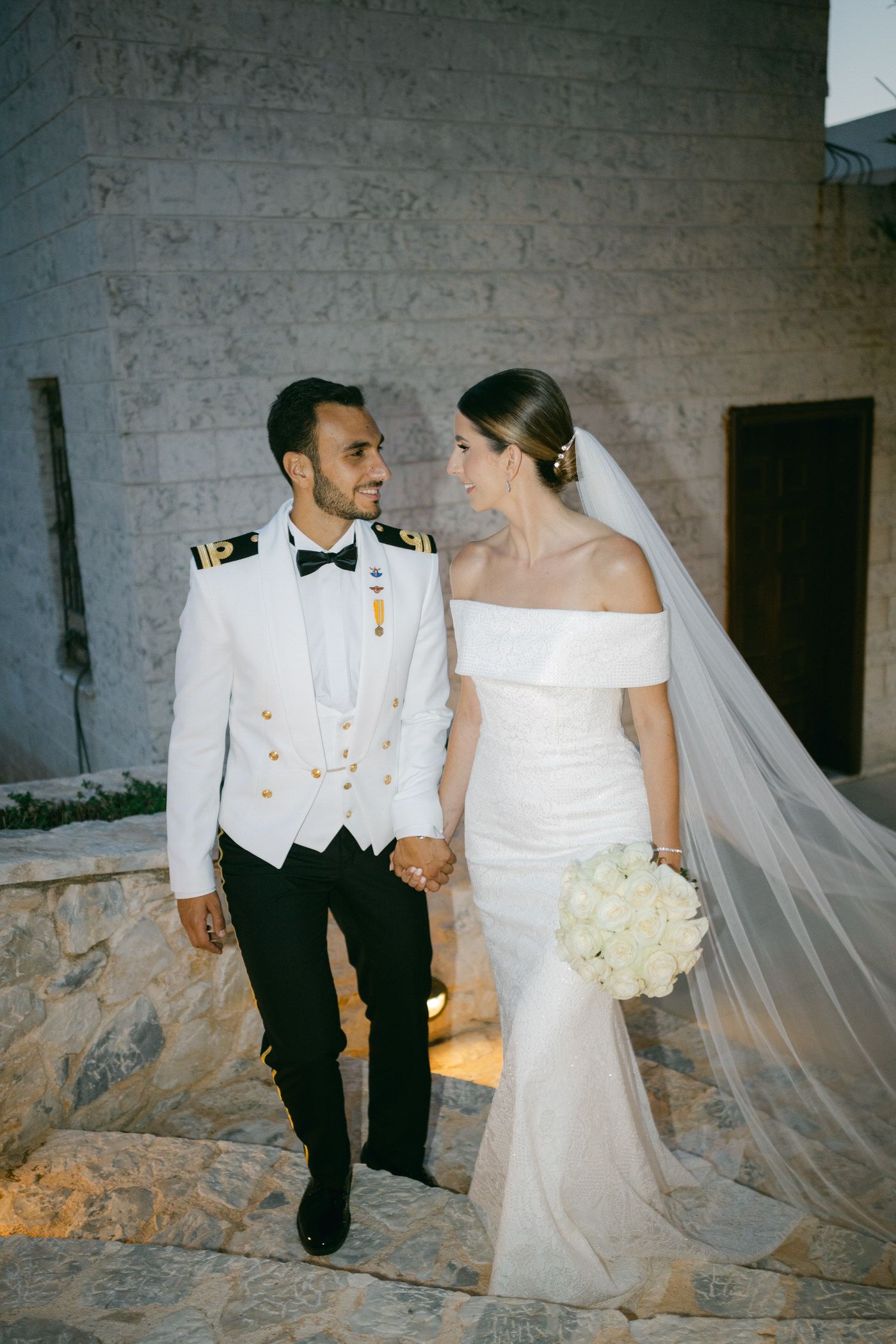 A bride in an off-shoulder gown and groom in naval uniform hold hands while walking up stone steps during their wedding.