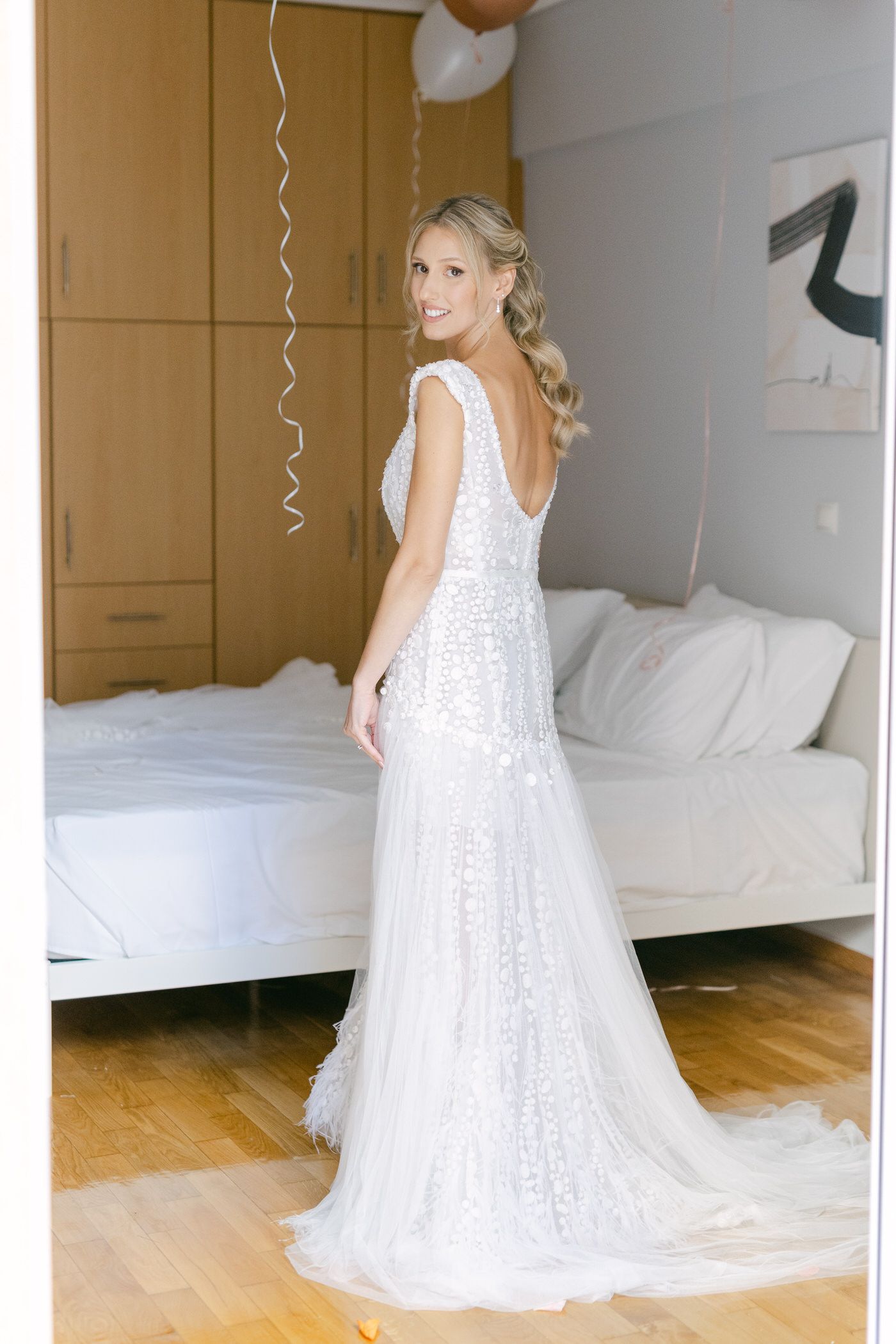 A bride wearing a flowing white lace wedding gown with cap sleeves stands in a bedroom looking over her shoulder.