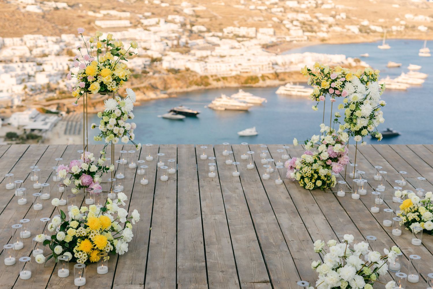 Floral arrangements with white and yellow blooms decorate a wooden deck overlooking a scenic myconos nammos view.
