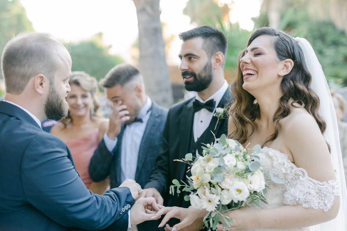 Wedding celebration with laughter and joy during outdoor ceremony as couple exchanges rings while holding white bouquet.