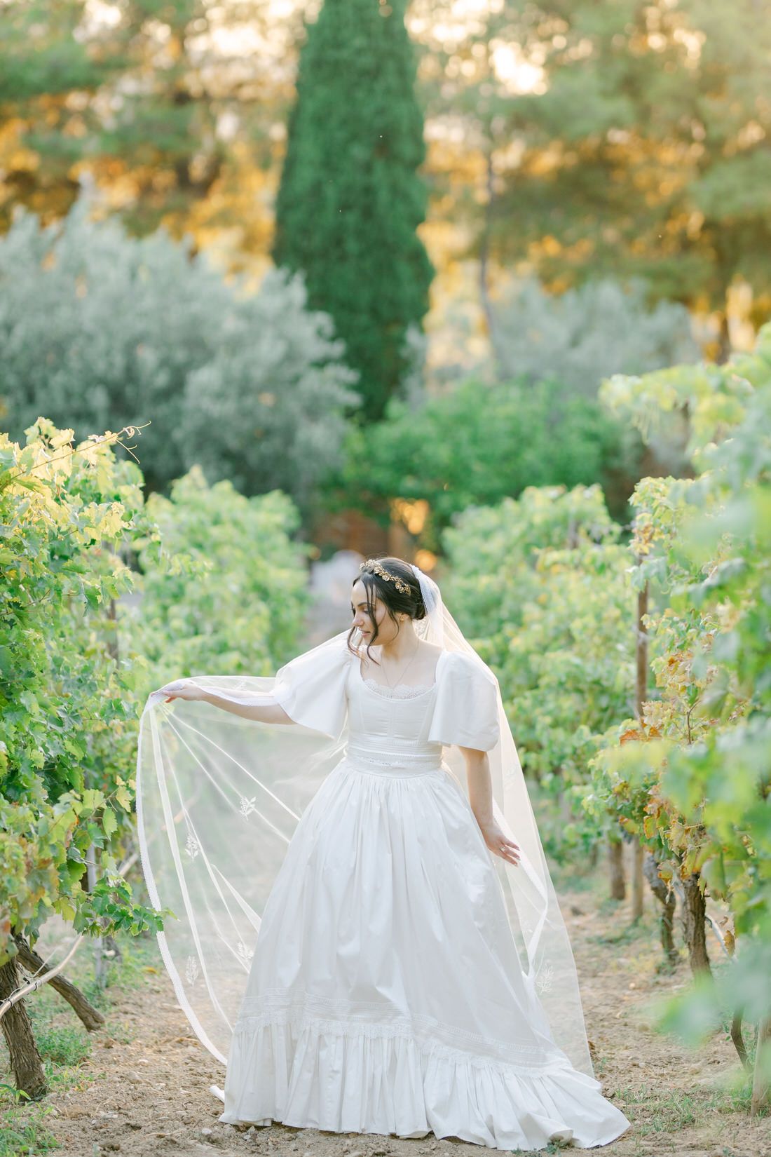 A white wedding dress flows elegantly in a vineyard garden setting with cypress trees and golden sunlight.