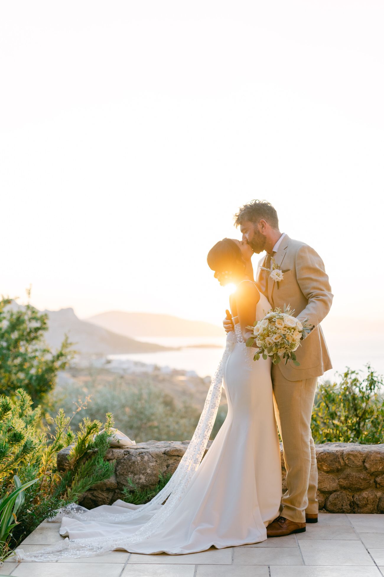 A romantic sunset wedding photo of a couple embracing on a stone terrace overlooking mountains.