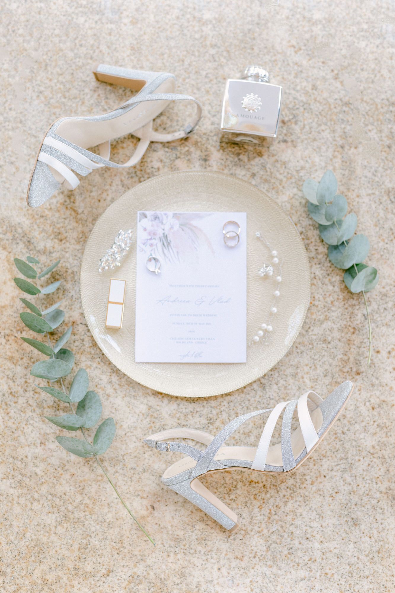 Wedding details flatlay with elegant white shoes, invitation, perfume bottle and eucalyptus sprigs on beige background.