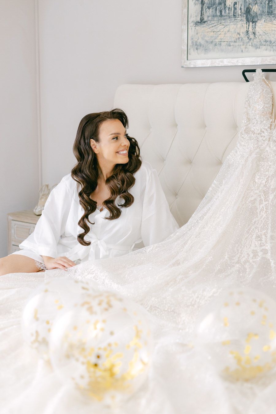Bride in white robe admires wedding dress displayed on tufted sofa.