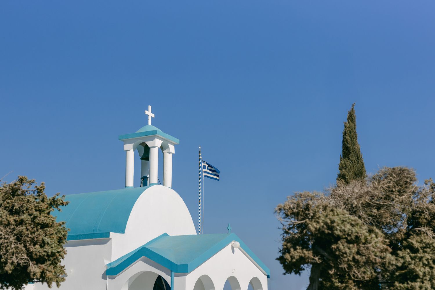 Traditional white Greek Orthodox church with turquoise dome and bell tower against bright blue sky in Cyprus.