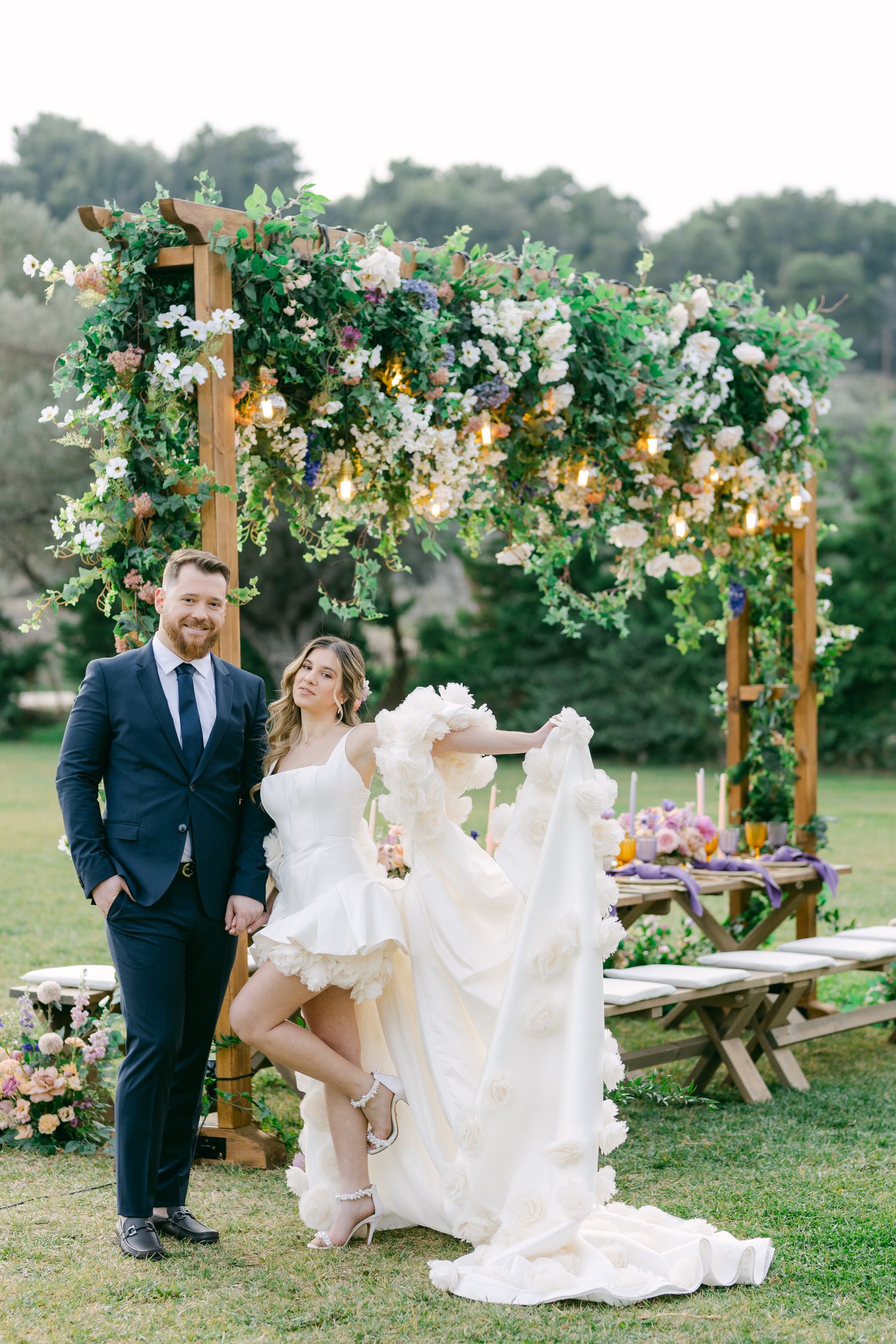 A romantic wedding scene with couple under a lush floral arch with white and green florals in an outdoor garden setting.