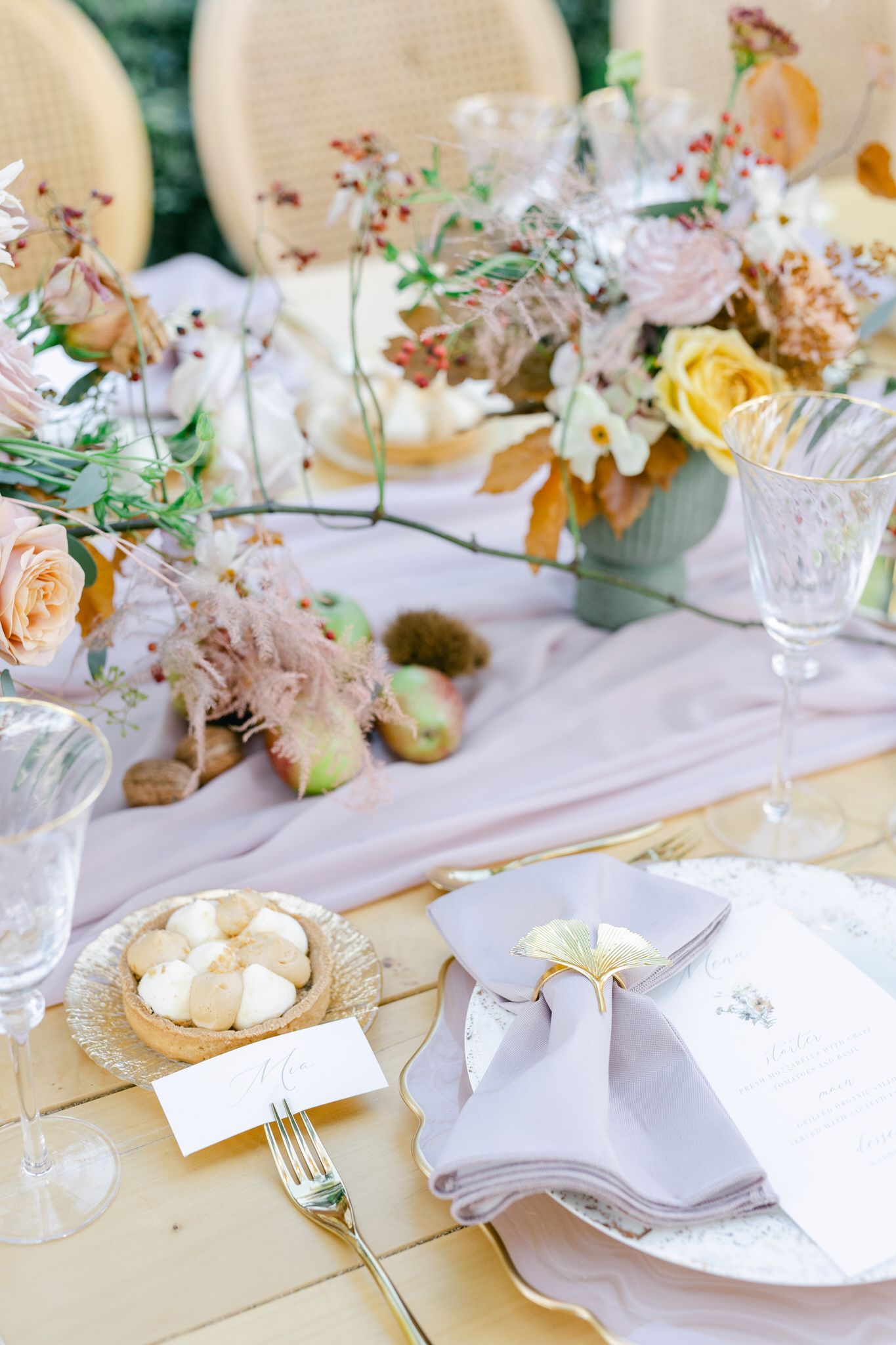 Elegant table setting with soft yellow and pink floral centerpiece, crystal glasses, and gray linen napkins on pale wood.