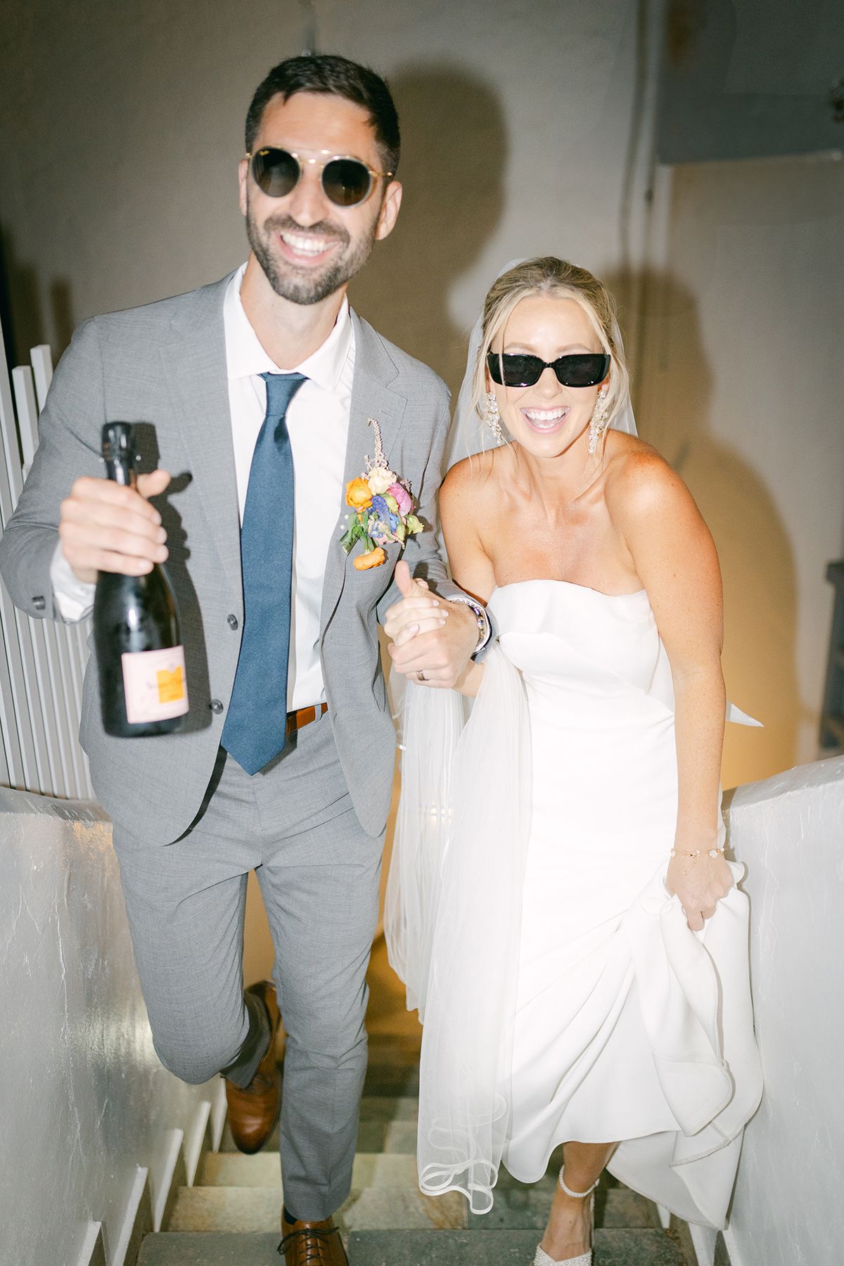 A bride and groom wearing sunglasses walk together holding champagne and flowers in a white stairwell.
