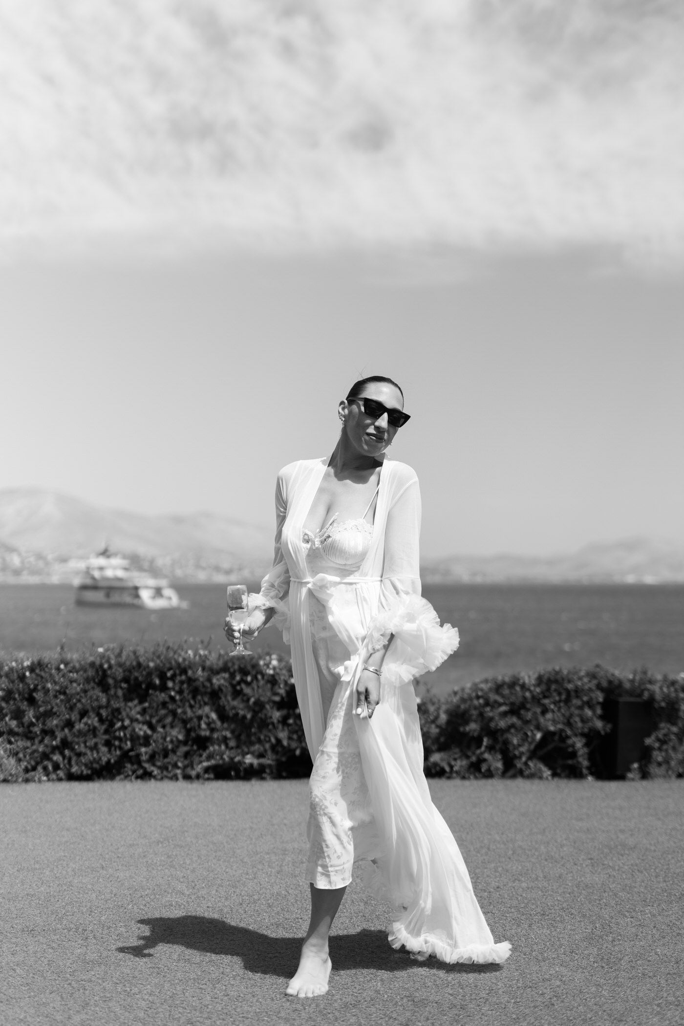 Black and white photo of someone in a flowing white dress and sunglasses posing barefoot on grass with water and hills behind.