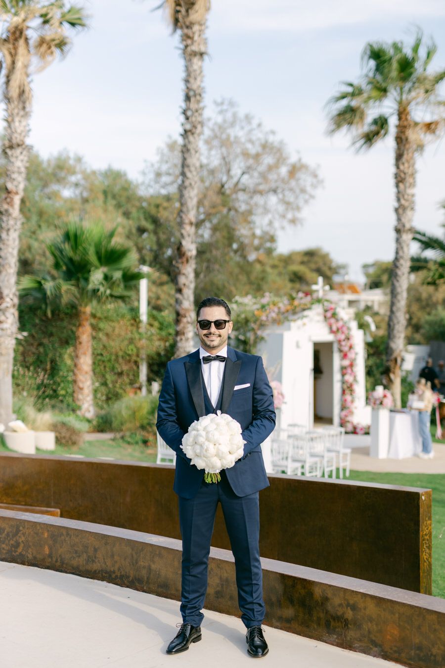 Groom in navy tuxedo holding white bouquet at tropical wedding venue with palm trees.