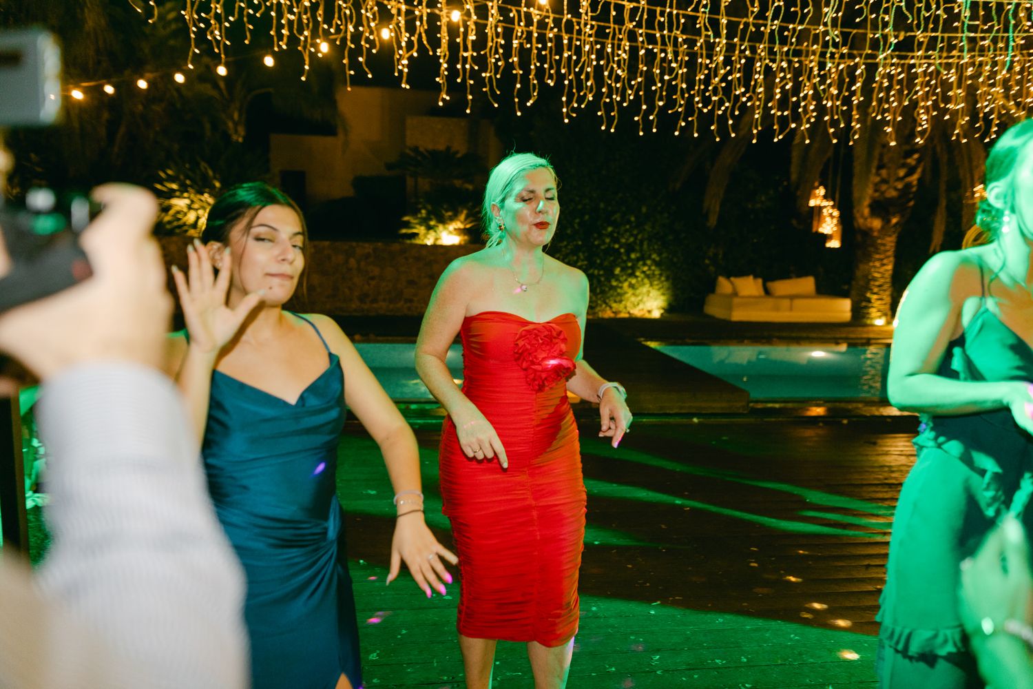 People dancing at a party under decorative string lights with a person in a red dress standing out in the center.
