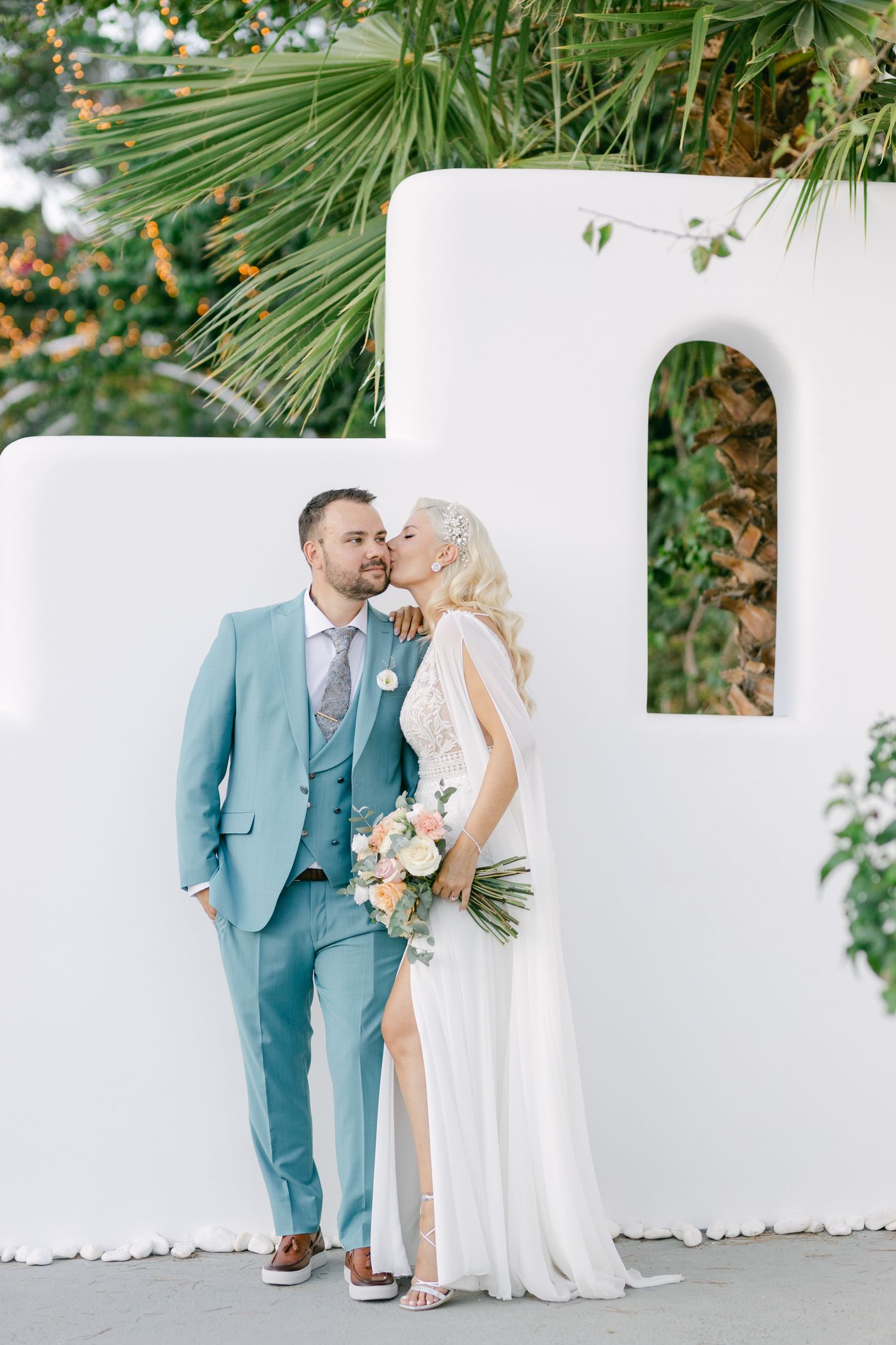 Romantic wedding couple in elegant attire pose against white Mediterranean-style wall with palm fronds overhead.
