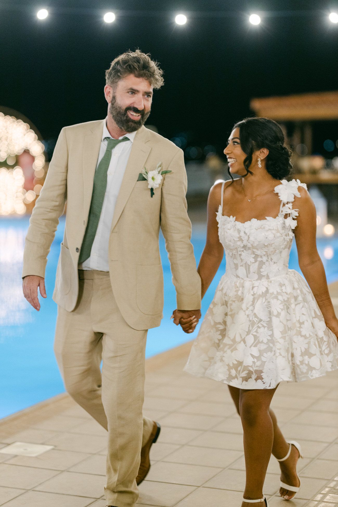 A couple walks hand in hand at night by a pool in wedding attire.