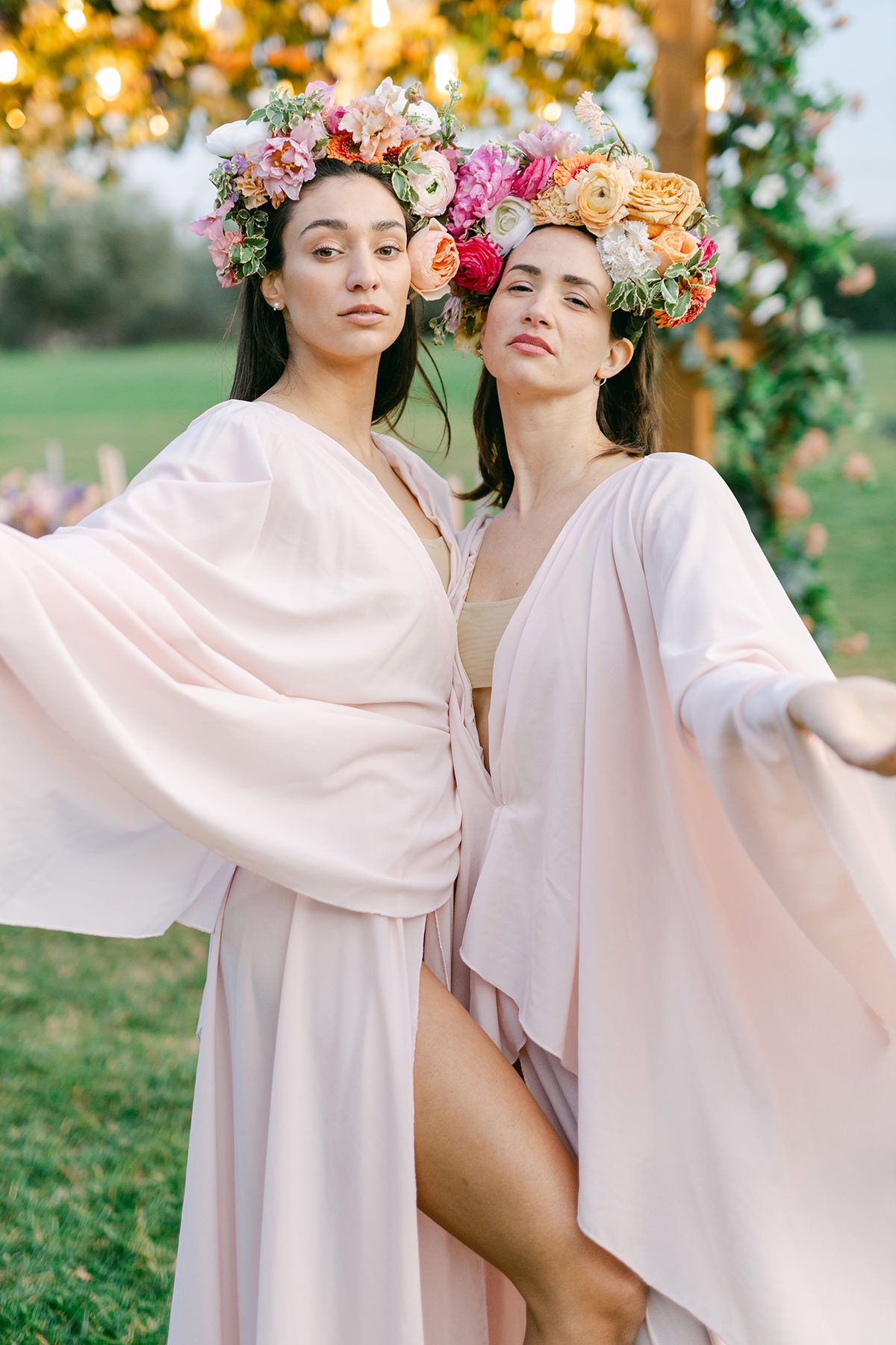 Two figures in flowing blush-colored robes pose together outdoors wearing floral crowns of pink and peach blooms.