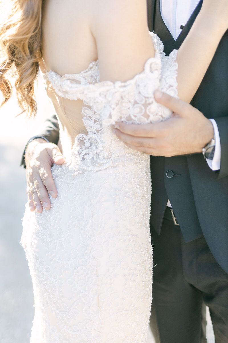 Close-up shot of bride in white lace gown being held by groom in traditional black tuxedo during wedding ceremony.
